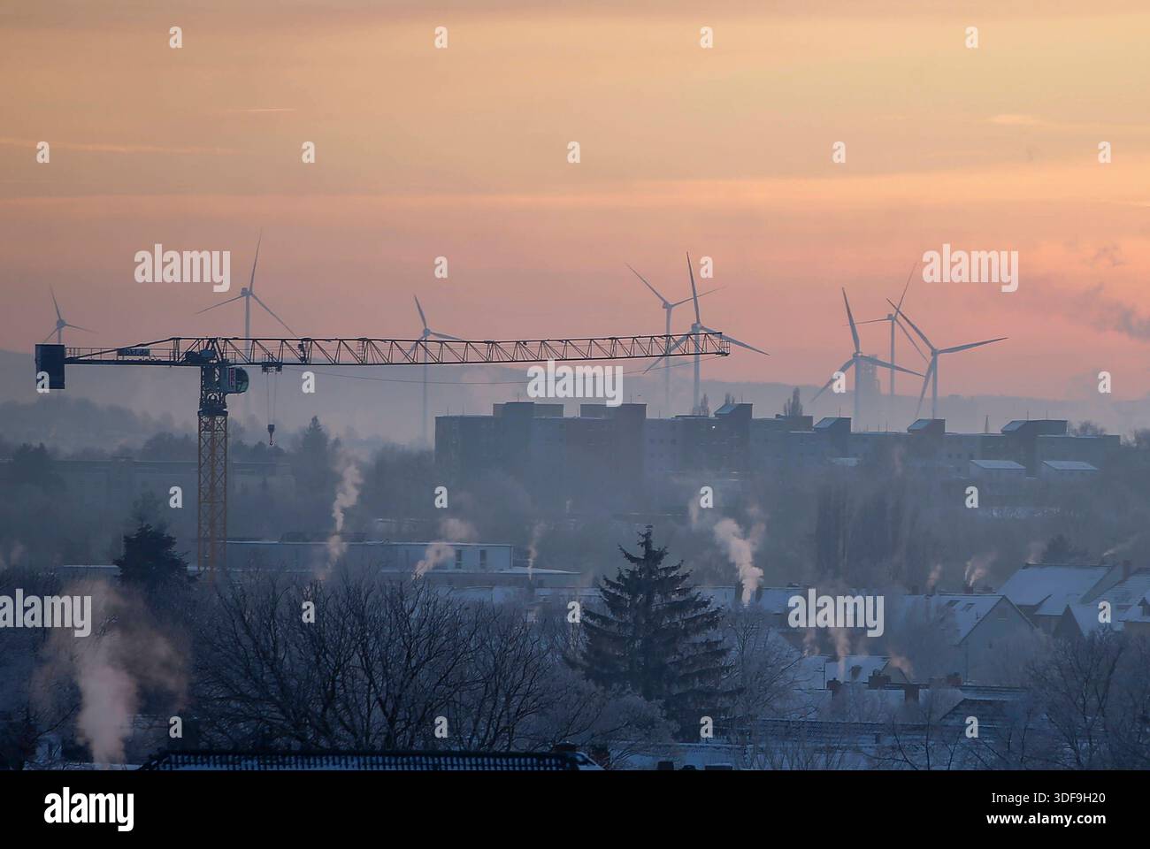 **Braunschweig, Deutschland, Europa.** Winterliche Stadtlandschaft in den frühen Morgenstunden. Baukran im Vordergrund über verschneiten Wohngebäuden, im Hintergrund mehrere Windkraftanlagen auf einem Höhenzug. Aufsteigender Dampf aus Schornsteinen, Nebel und Dunst in der kalten Luft. Pastellfarbener Himmel bei Sonnenaufgang, eingeschränkte Sicht durch Inversionslage. Erhöhte Perspektive, weite totale, keine Personen erkennbar. Braunschweig, Winter, Stadtansicht, Sonnenaufgang, Baukran, Windkraft, energie, Infrastruktur, Kälte, Dunst, Wetter, Klima, Umwelt, Gesellschaft, Symbolfoto, notizie. *** Foto Stock