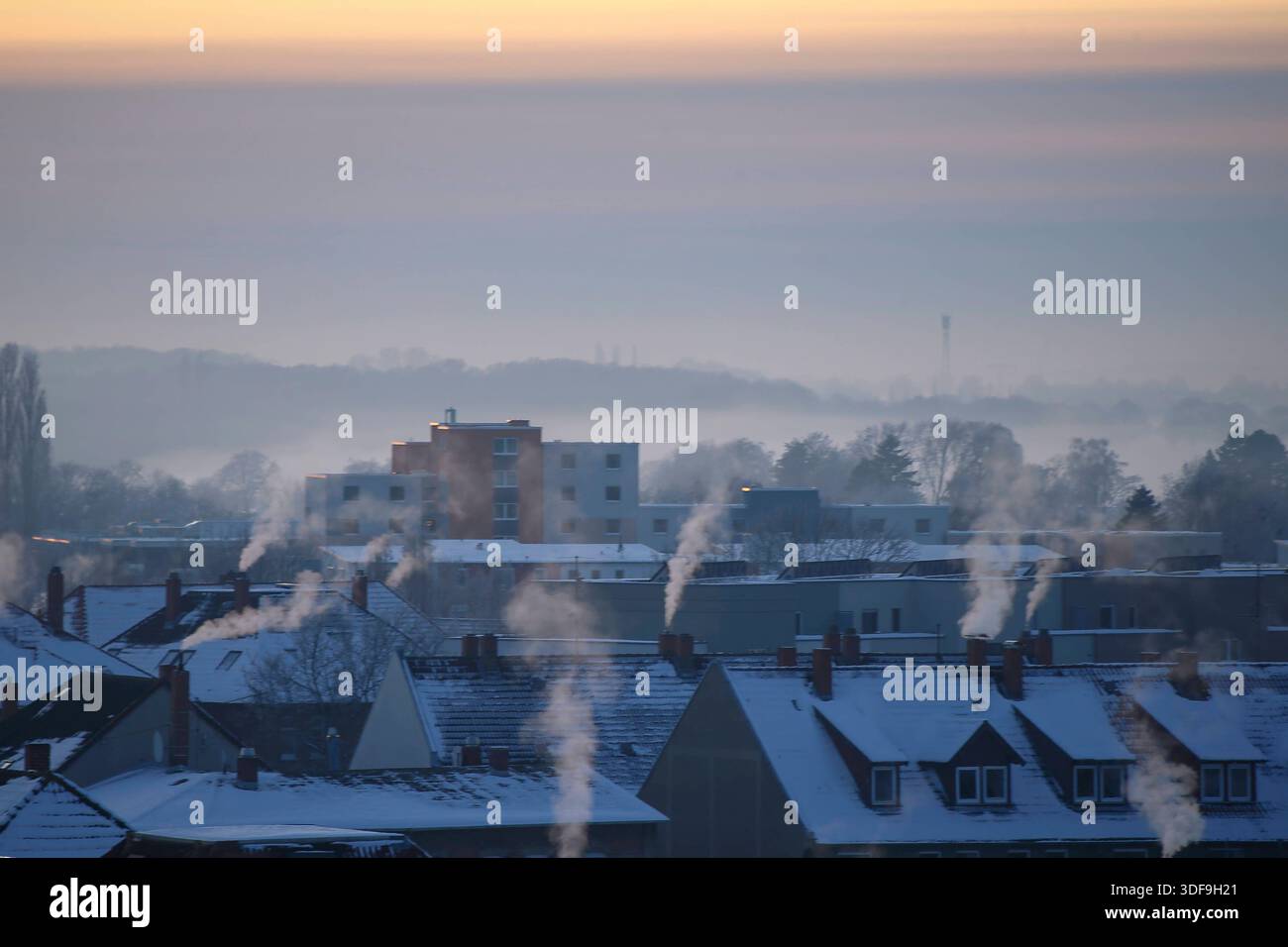 **Braunschweig, Deutschland, Europa.** Winterliche Wohnbebauung in den frühen Morgenstunden. Verschneite Dächer mit zahlreichen Schornsteinen, aus denen Wasserdampf aufsteigt. Dunst und Nebel liegen über dem Stadtgebiet, im Hintergrund Baumreihen und niedrigere Gebäude, teilweise eingeschränkte Sicht. Kühle Farbstimmung, ruhige Wetterlage mit Inversionsschicht. Erhöhte Perspektive, weite totale, keine Personen sichtbar. Braunschweig, Winter, Kälte, Nebel, Dunst, Wohngebiet, Schornsteine, Heizperiode, Stadt, Wetter, Umwelt, Klima, Gesellschaft, Symbolfoto, News. *** Brunswick, Germania, Europa W. Foto Stock