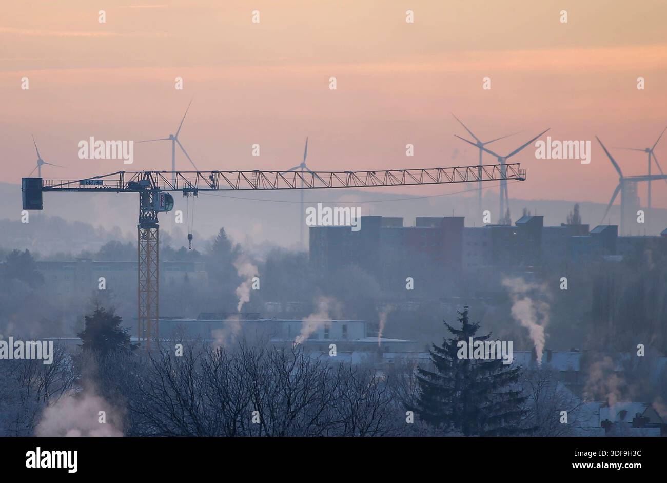 **Braunschweig, Deutschland, Europa.** Winterliche Stadtlandschaft in den frühen Morgenstunden. Baukran im Vordergrund über verschneiten Wohngebäuden, im Hintergrund mehrere Windkraftanlagen auf einem Höhenzug. Aufsteigender Dampf aus Schornsteinen, Nebel und Dunst in der kalten Luft. Pastellfarbener Himmel bei Sonnenaufgang, eingeschränkte Sicht durch Inversionslage. Erhöhte Perspektive, weite totale, keine Personen erkennbar. Braunschweig, Winter, Stadtansicht, Sonnenaufgang, Baukran, Windkraft, energie, Infrastruktur, Kälte, Dunst, Wetter, Klima, Umwelt, Gesellschaft, Symbolfoto, notizie. *** Foto Stock