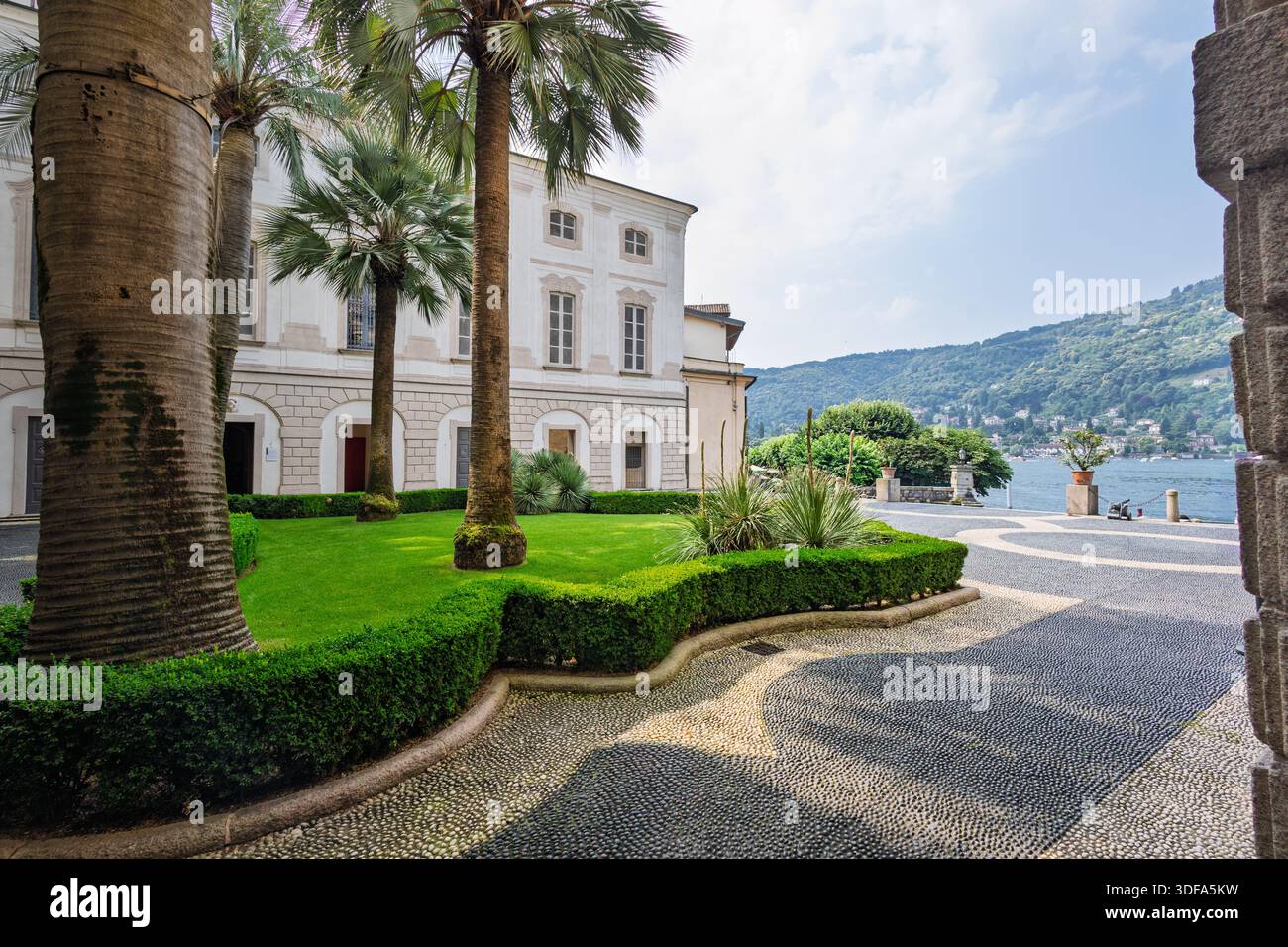 Un grande arco incornicia un tranquillo cortile caratterizzato da un audace mosaico di ciottoli, un edificio classico bianco e una vegetazione ben curata. Architettura senza tempo Foto Stock