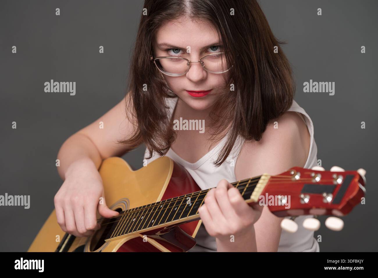 Donna chitarrista musicista rock che esegue un pezzo di musica su una classica chitarra acustica a sei corde d'acciaio. Giovane musicista adulto che indossa occhiali che sembrano ca Foto Stock