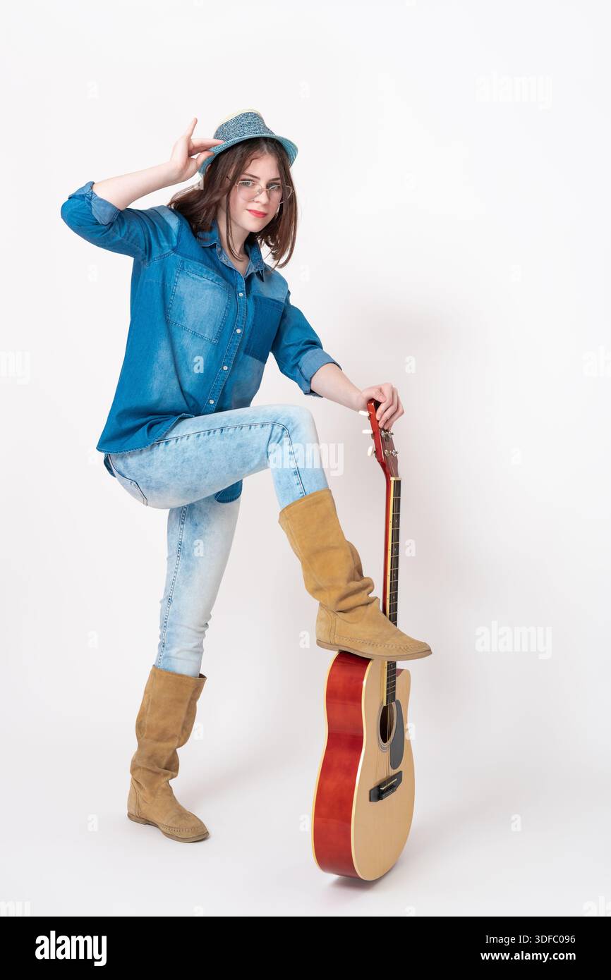 Chitarrista donna e chitarra in piedi davanti, posiziona una gamba drappeggiata sulla chitarra e guarda la fotocamera. Musicista donna che indossa occhiali da vista, d Foto Stock