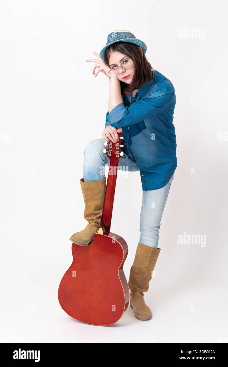 Bella chitarrista con la chitarra in piedi davanti. Posiziona una gamba drappeggiata sulla chitarra e guarda la telecamera. Musicista in occhiali, Foto Stock