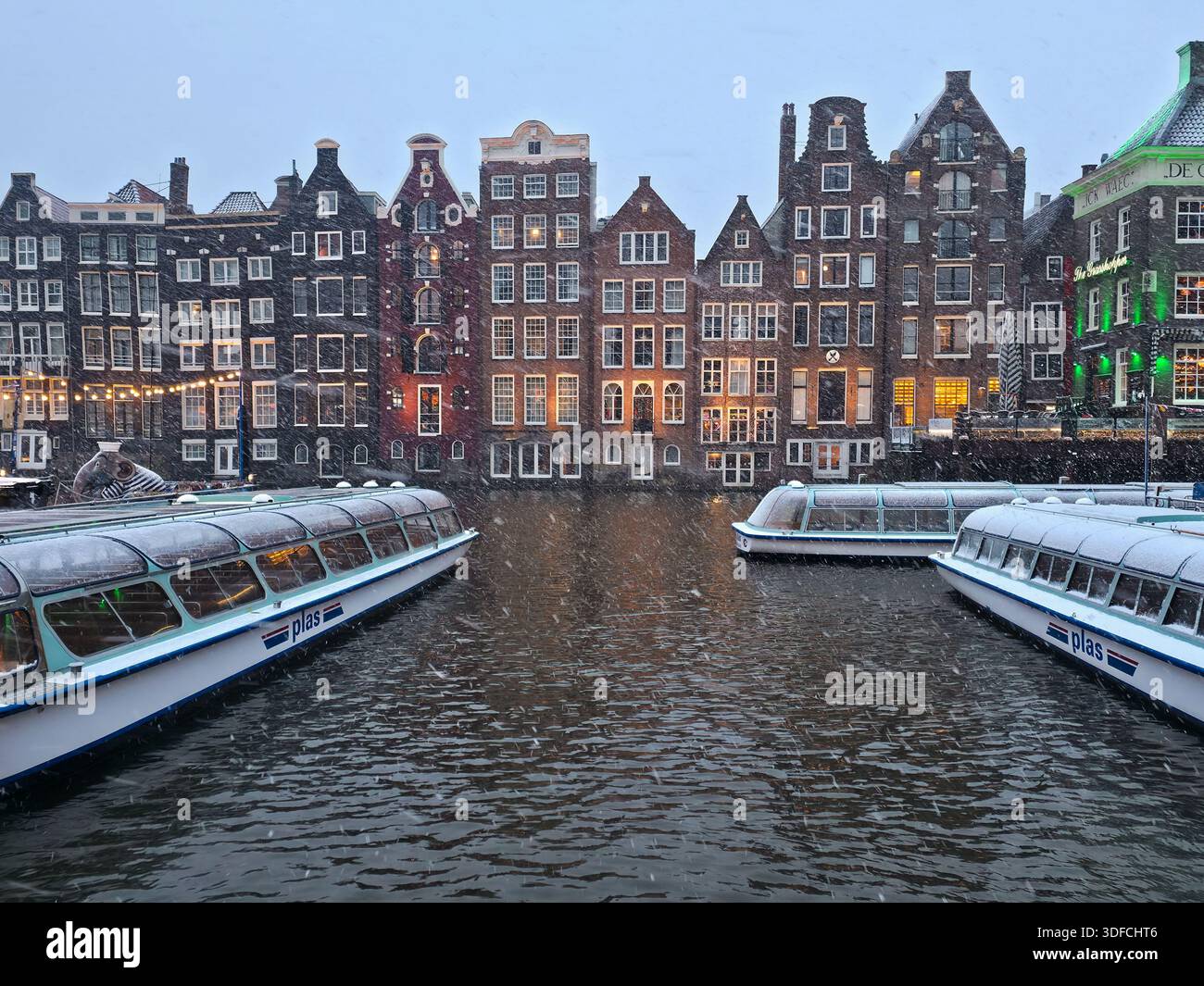 La neve cade sulle storiche case sui canali di Amsterdam all'ora blu, le calde luci che si riflettono sull'acqua mentre una casa galleggiante passa davanti alle iconiche facciate olandesi Foto Stock