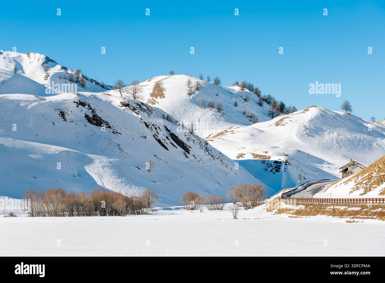 Vista panoramica delle piste innevate, una strada tortuosa e un lago ghiacciato al passo della Maddalena, sotto un cielo azzurro. Foto Stock