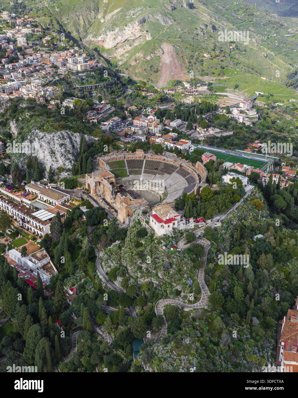 Vista aerea dell'antico Teatro Antico, immerso nel verde e nei sentieri tortuosi, contrasta con i tetti in terracotta della città, Taormina, S. Foto Stock