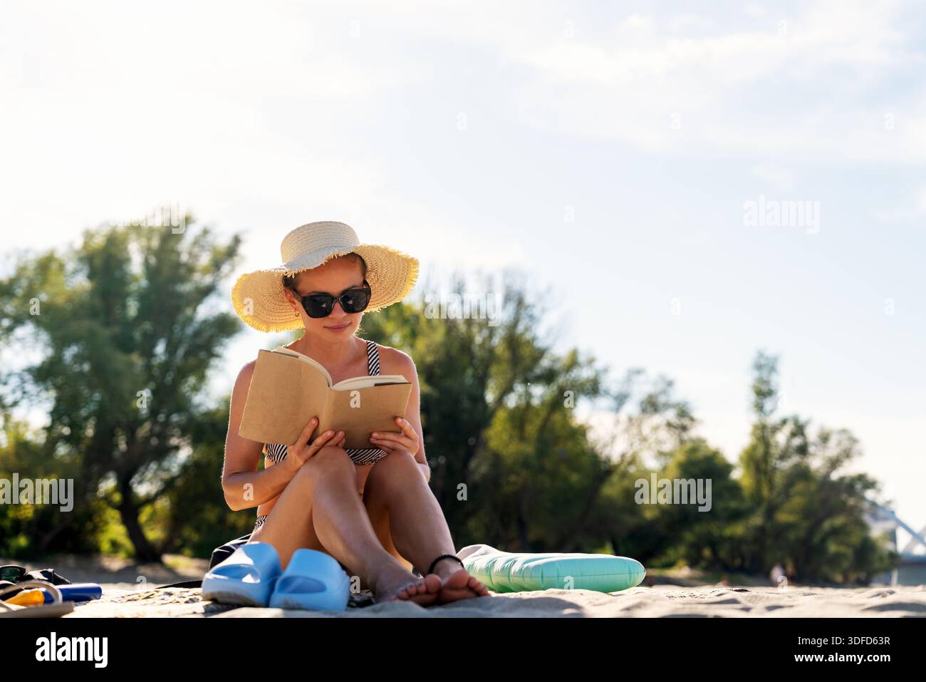 Donna con cappello e occhiali da sole che legge un libro sulla spiaggia in estate Foto Stock