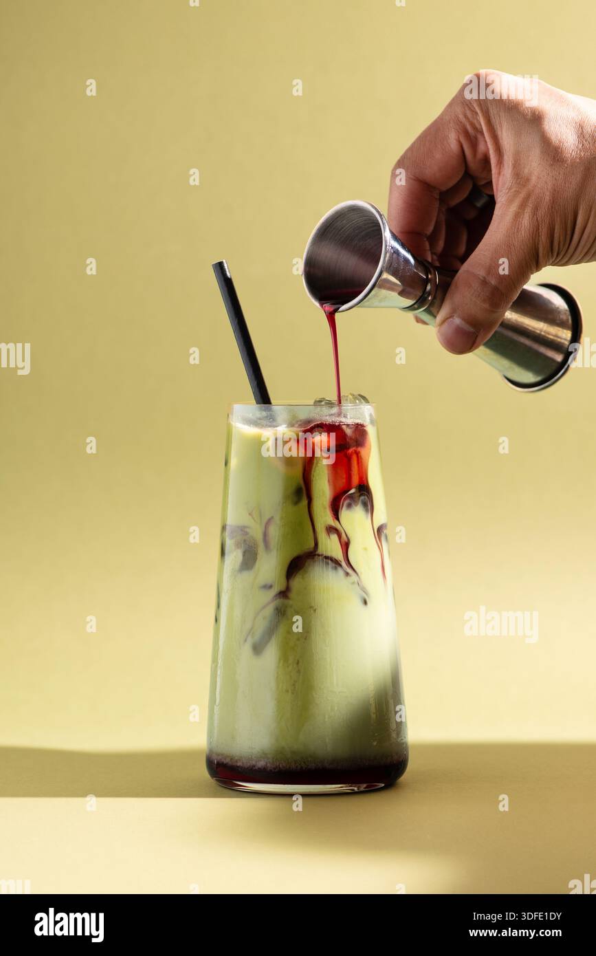 Bevanda matcha al lampone ghiacciata con sciroppo rosso versato da un jigger in un bicchiere trasparente. Bevanda a strati colorata. Foto Stock