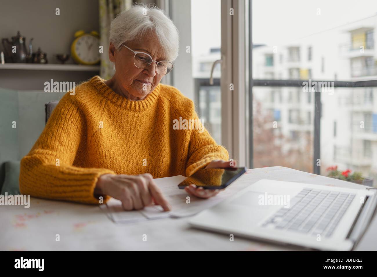 Donna anziana che tiene il telefono cellulare mentre legge documenti cartacei a casa Foto Stock