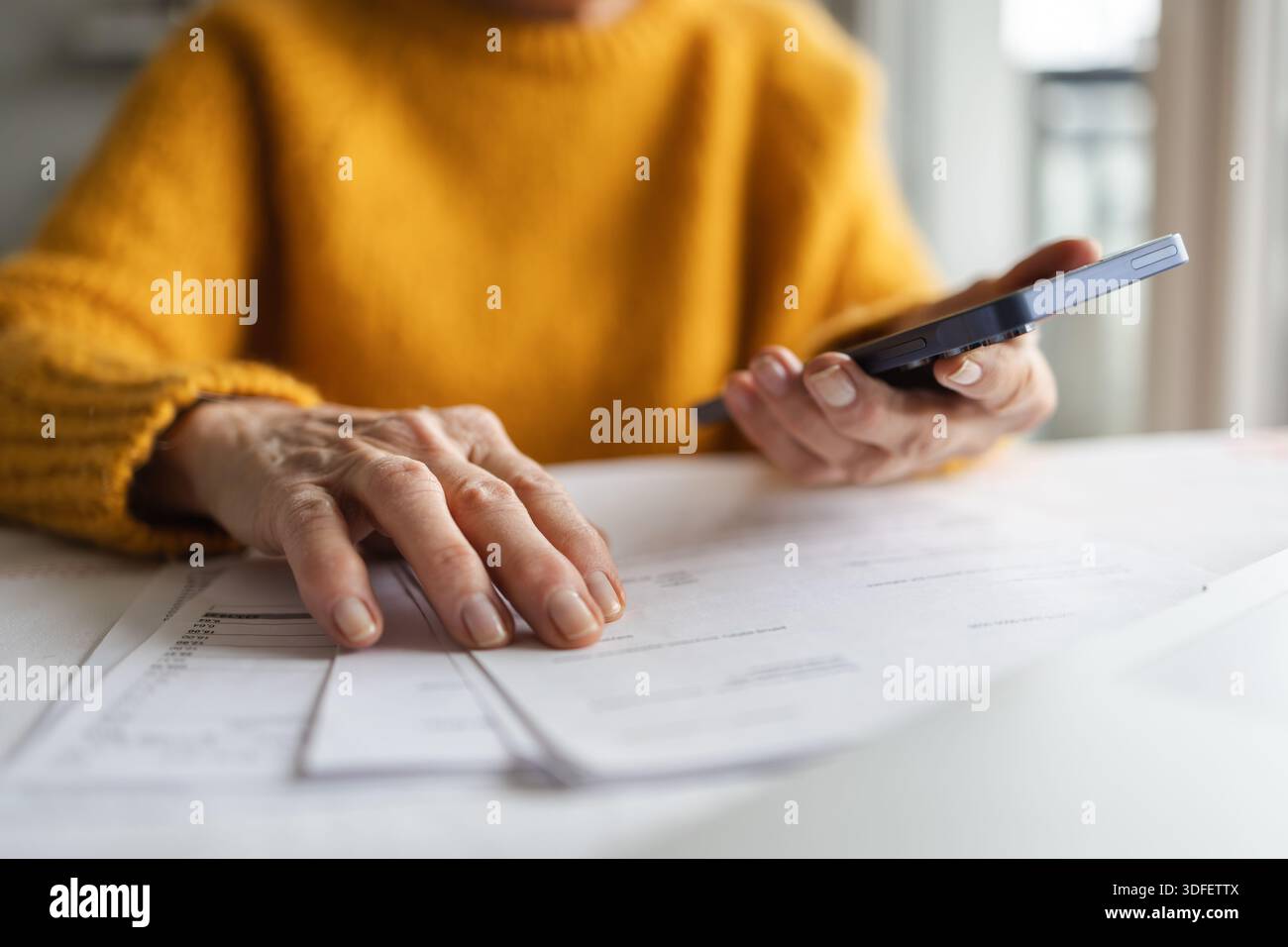 Donna anziana che tiene il telefono cellulare mentre legge documenti cartacei a casa Foto Stock