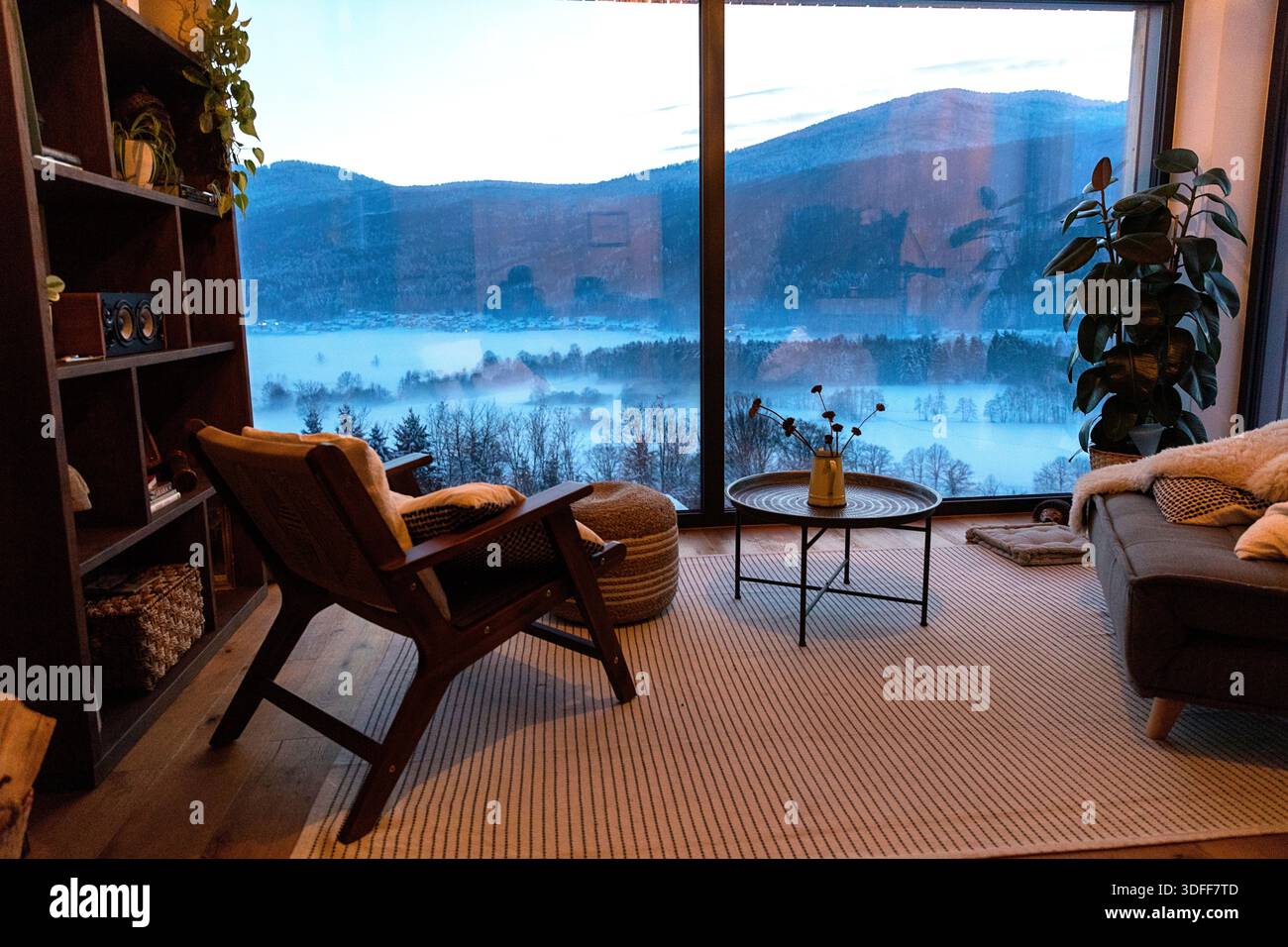 Intimo soggiorno interno in stile scandinavo con finestra panoramica con mobili minimalisti, piante e vista panoramica sulla campagna invernale Foto Stock
