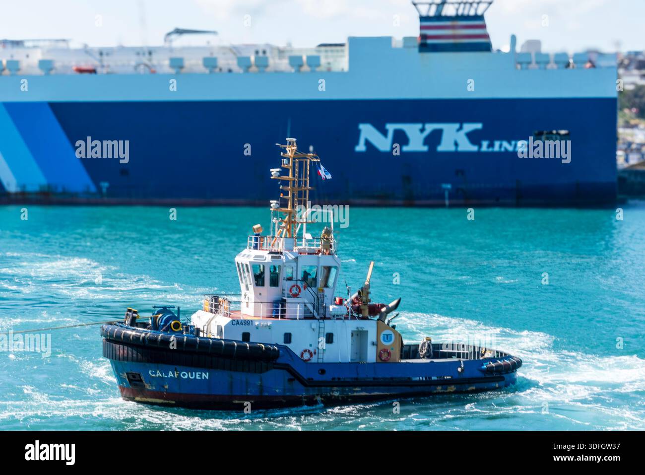 Rimorchiatore del porto che passa per le calme acque del porto con la nave portaerei NYK ormeggiata sullo sfondo. Foto Stock