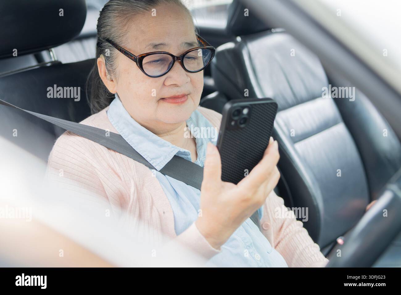 Donna anziana sorridente che tiene in mano lo smartphone mentre è seduta in auto, donna matura che guida l'auto e utilizza il telefono. Concetto di stile di vita pensionistico. Foto Stock