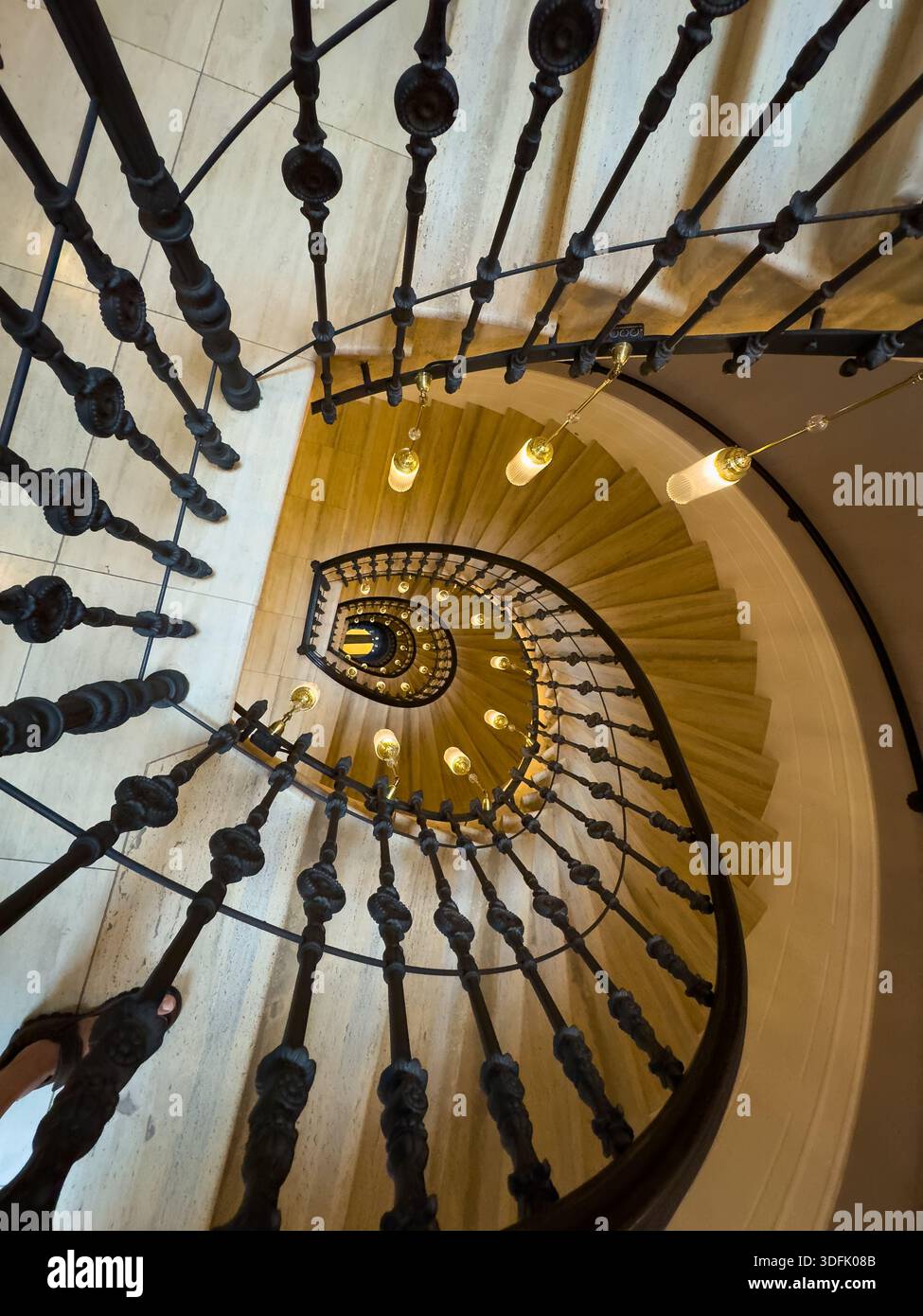 Le scale in legno a spirale si curvano verso il basso sotto i balaustri di ferro ornati illuminati da luci a sospensione vintage che creano un punto focale architettonico spettacolare. Foto Stock