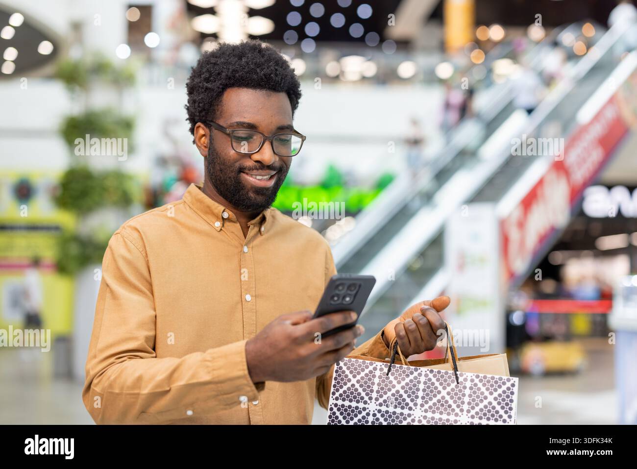 Giovane felice che indossa occhiali con una borsa della spesa di marca, sorridendo mentre controlla lo smartphone per ricevere notifiche, messaggi o acquisti online Foto Stock