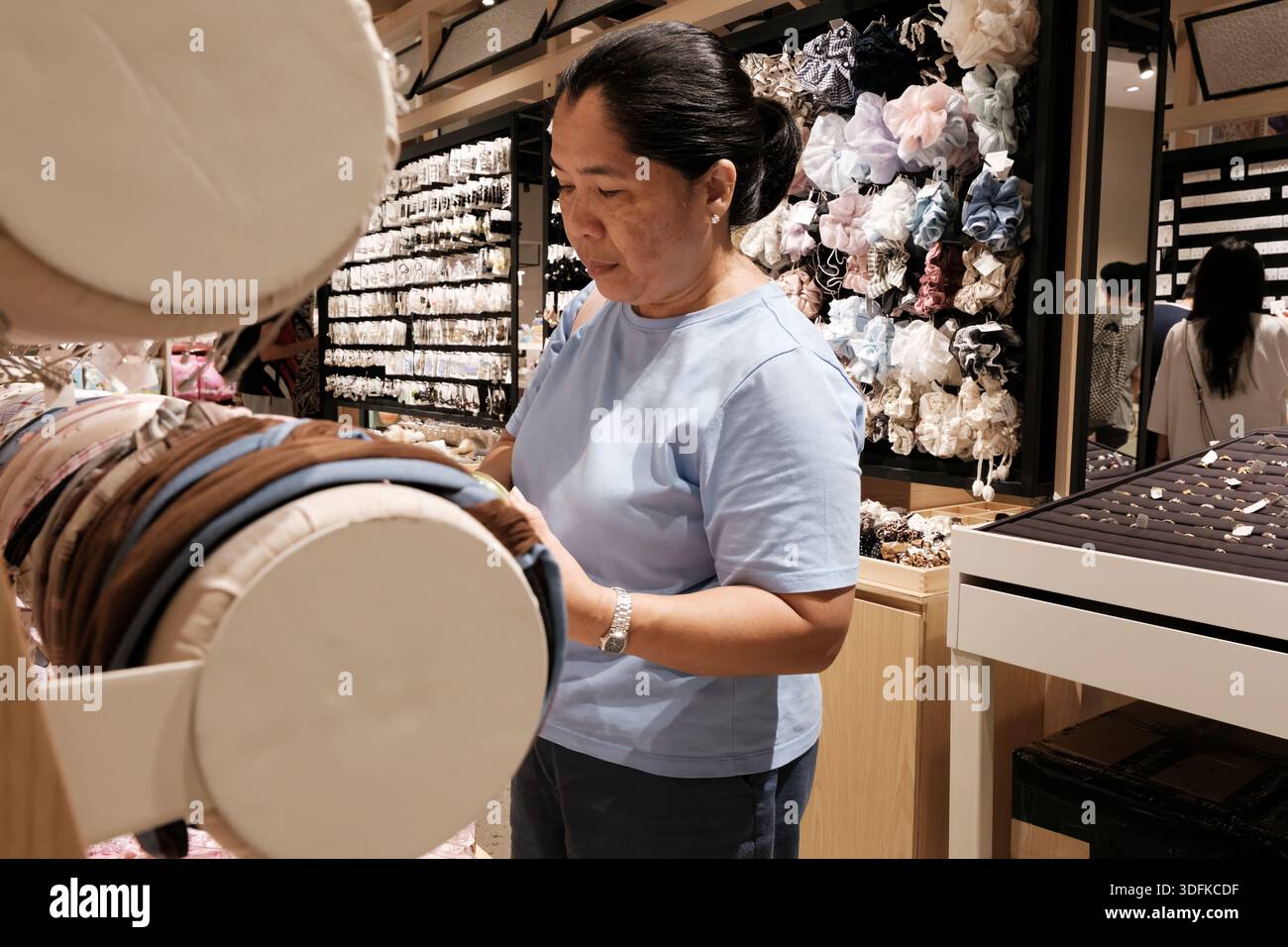 Una donna si trova in un negozio che guarda gli accessori per capelli. Il negozio ha molti oggetti colorati in mostra e persone che navigano nelle vicinanze. E' giorno. Foto Stock