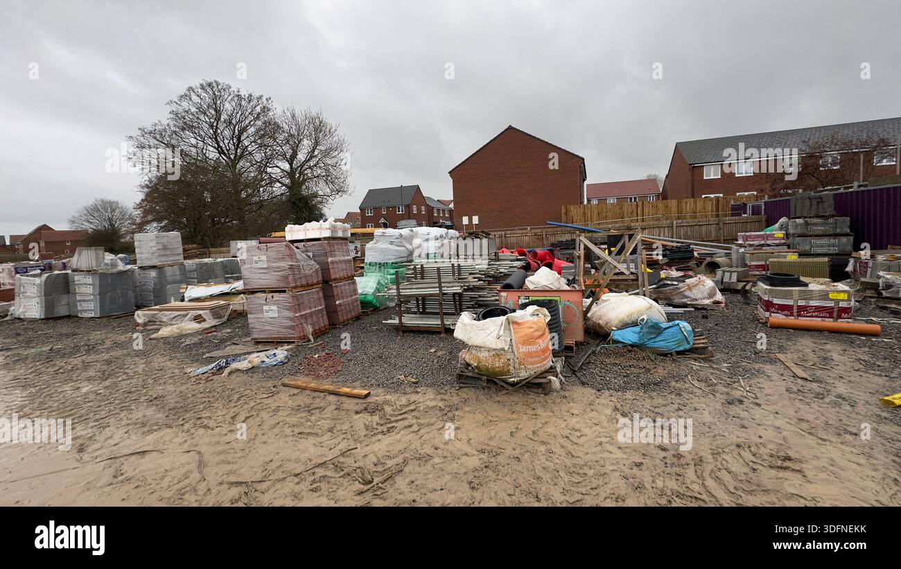 Un cantiere con pile di materiali da costruzione, tra cui mattoni e blocchi di cemento, sotto un cielo nuvoloso. Sono visibili terreni fangosi e alberi. Foto Stock