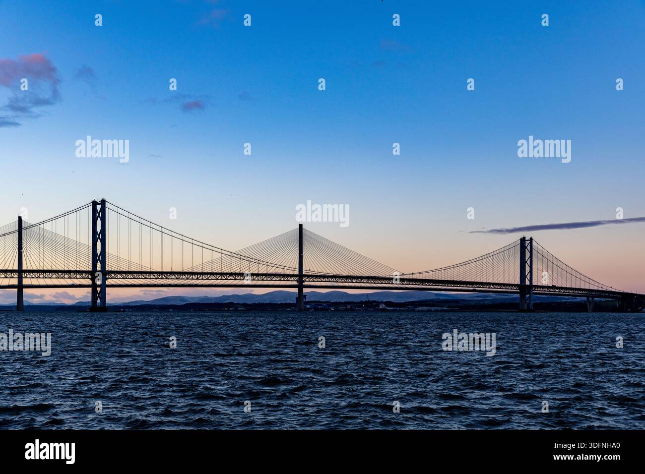 Un ampio ponte sospeso si estende su un corpo d'acqua scuro e instabile sotto un cielo digradante di blu e arance morbide, con colline lontane che si stagliano. Foto Stock
