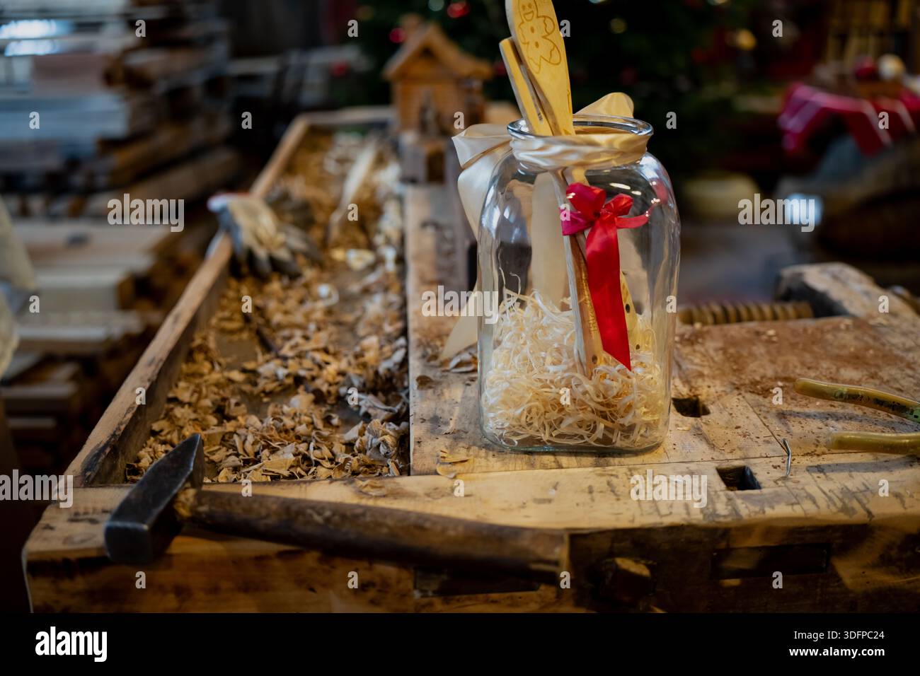 Laboratorio rustico di natura morta che mostra un regalo chiaro in vaso con nastri e ornamenti in legno, seduto su un vecchio banco da lavoro con trucioli di legno, martello e mazze artigianali Foto Stock