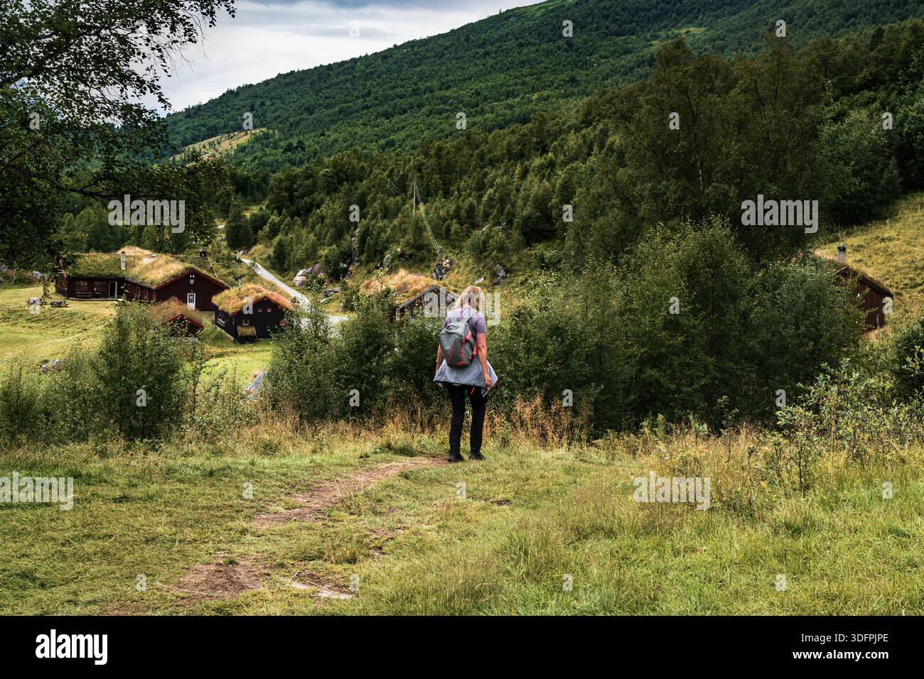 Una donna in spalla cammina lungo un sentiero escursionistico nella valle di Innerdalen. Le tradizionali cabine norvegesi in legno sul tetto sono visibili tra gli alberi verdi delle colline Foto Stock