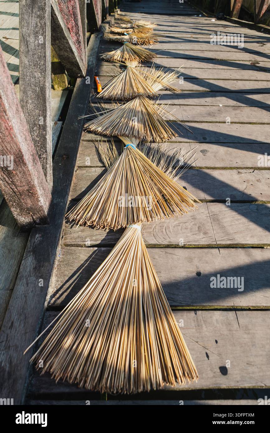 Diversi fasci di paglia naturale o fronde di palma, preparati per la preparazione di spazzatrici tradizionali (sapu lidi), sono disposti in fila per asciugare su una p di legno illuminata dal sole Foto Stock