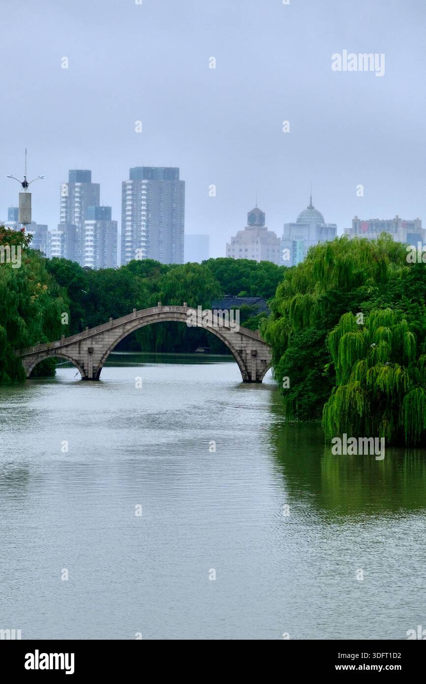 Un contrasto sorprendente catturato in una città cinese, caratterizzato da uno storico ponte ad arco in pietra circondato da lussureggianti alberi verdi di salice sopra un fiume tranquillo Foto Stock