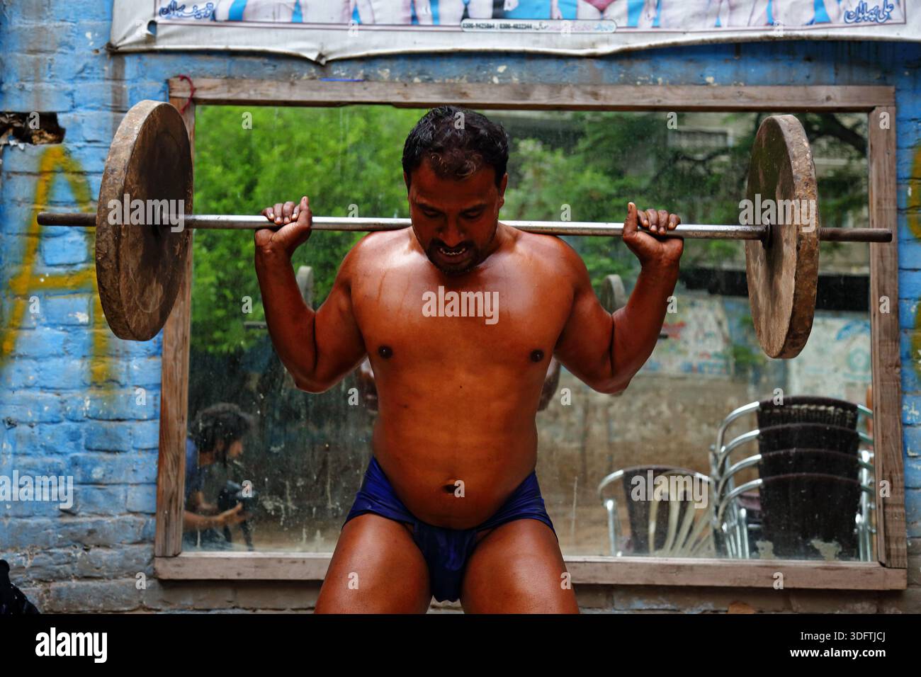 Lahore, Pakistan - 24 dicembre 2025: Vista di un determinato sollevatore di pesi in pantaloncini blu, che si sfidano sotto il barbell in una palestra all'aperto contro un vibrante Foto Stock