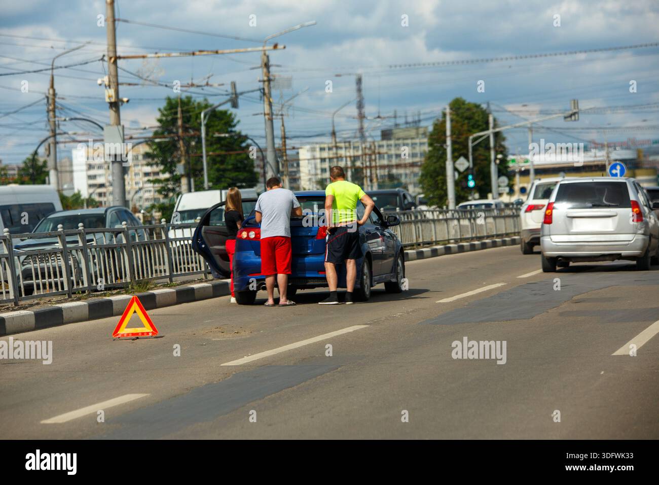 Tre giovani preoccupati, una donna e due uomini, esaminano un'auto blu su una strada dopo un incidente stradale durante il giorno. Foto Stock