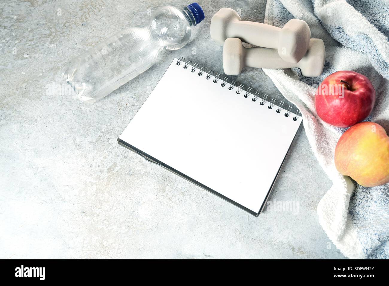 Quaderno ad anello vuoto per un elenco di obiettivi di fitness circondati da manubri, mele, bottiglia d'acqua e un asciugamano su pietra grigia, mockup per dimagrire e un hea Foto Stock