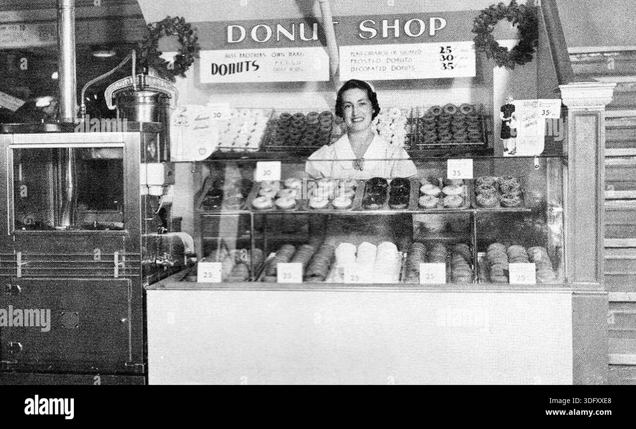 Questa fotografia mostra un negozio di ciambelle gestito da Hess Brothers ad Allentown, Pennsylvania, nel 1938. Riflette la vita urbana americana di tutti i giorni durante la tarda grande depressione, quando piccole aziende alimentari come i negozi di ciambelle servivano come spazi sociali accessibili per i clienti della classe lavoratrice e media. Negozi come questo facevano parte della crescente cultura dei consumatori americani degli anni '1930, offrendo prelibatezze economiche e sottolineando l'efficienza, la pulizia e la moderna presentazione al dettaglio. Foto Stock