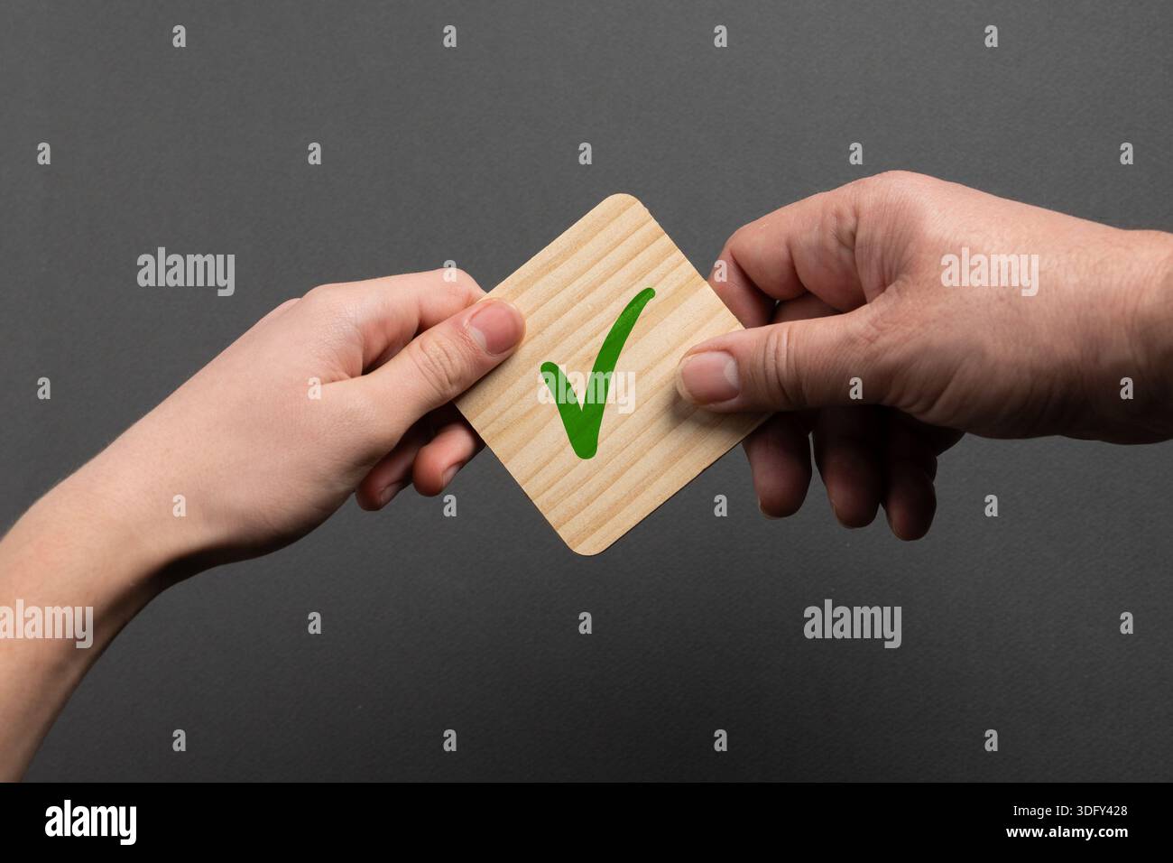 Primo piano di due mani con una carta di legno con un segno di spunta verde su sfondo grigio, che simboleggia l'approvazione, la conferma e la decisione positiva. Foto Stock