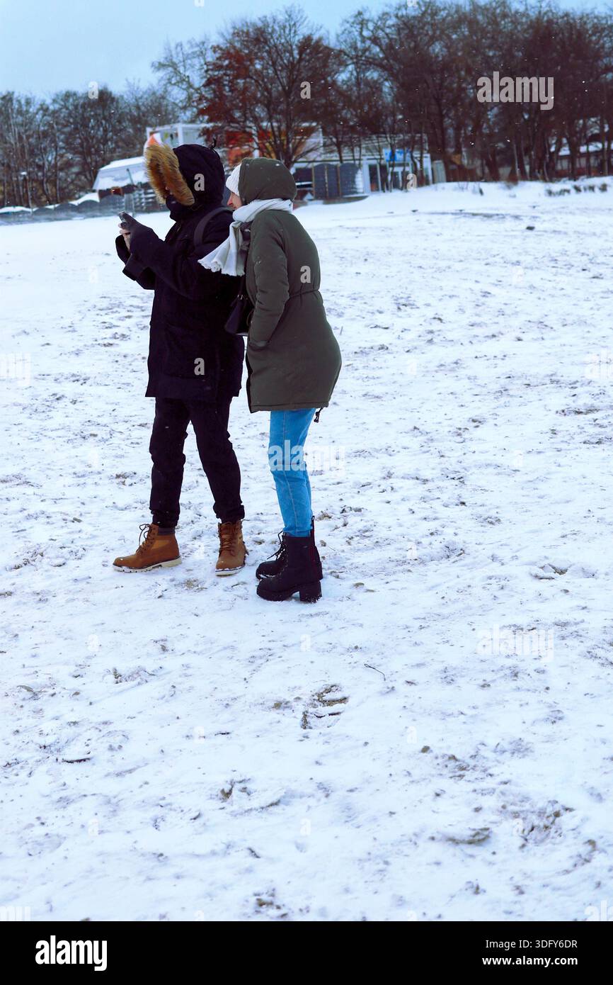 Due persone con cappotti invernali in piedi su una spiaggia innevata che guardano un cellulare Foto Stock