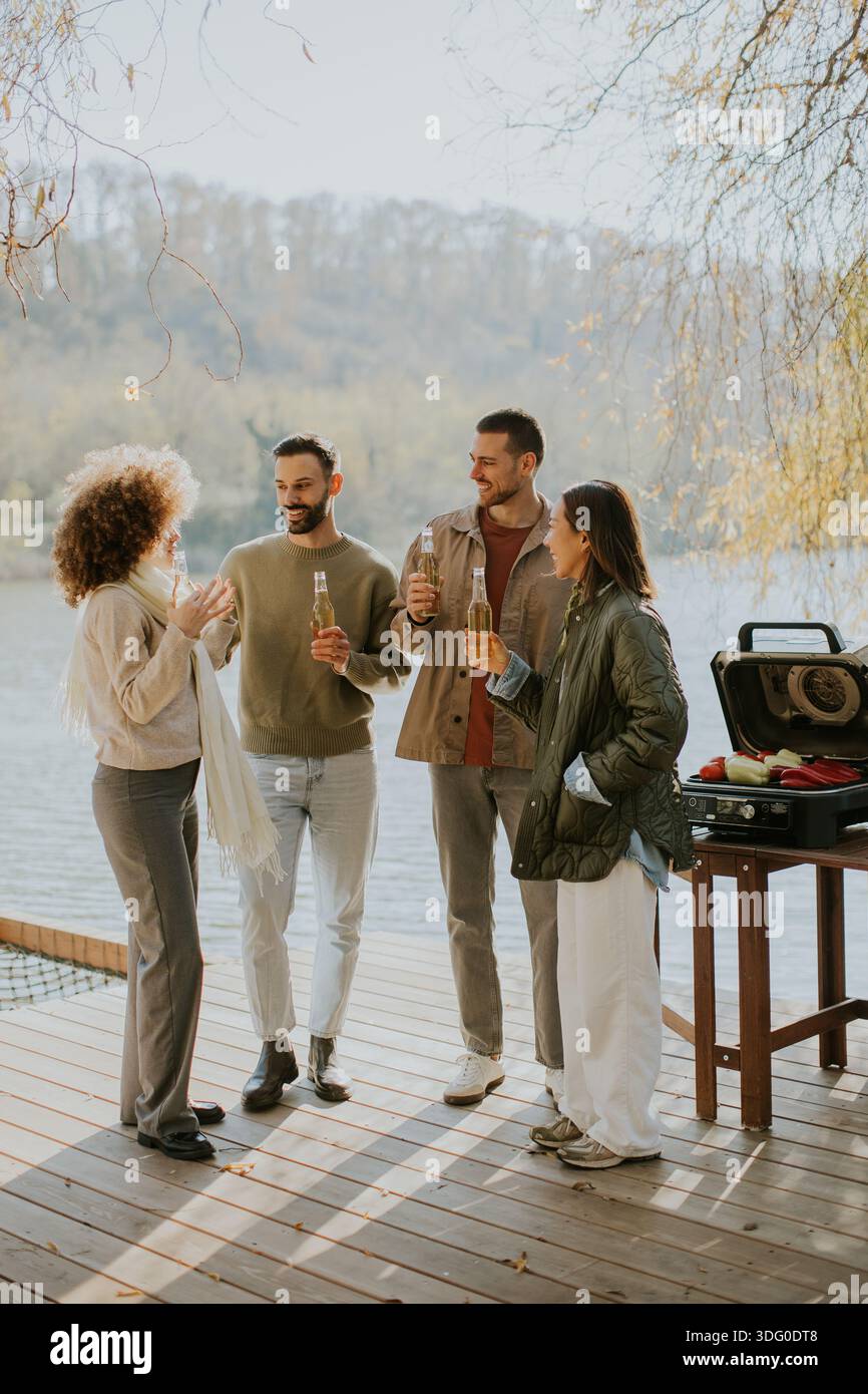 Quattro amici si riuniscono su un ponte di legno vicino all'acqua, condividendo bevande e risate mentre si godono la splendida giornata autunnale. Foto Stock