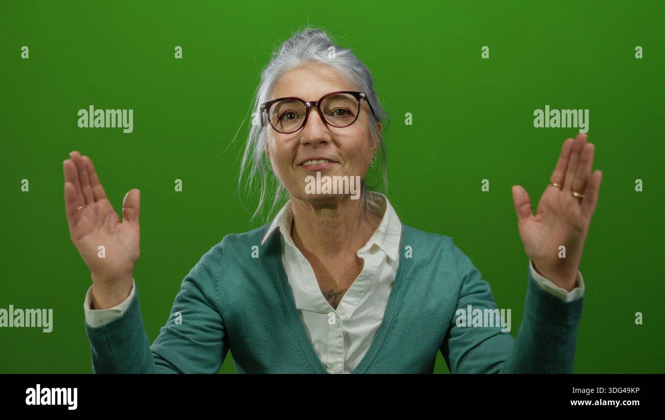 Donna anziana con capelli e occhiali grigi che gestiscono grandi dimensioni su una parete di sfondo verde isolata. Foto Stock
