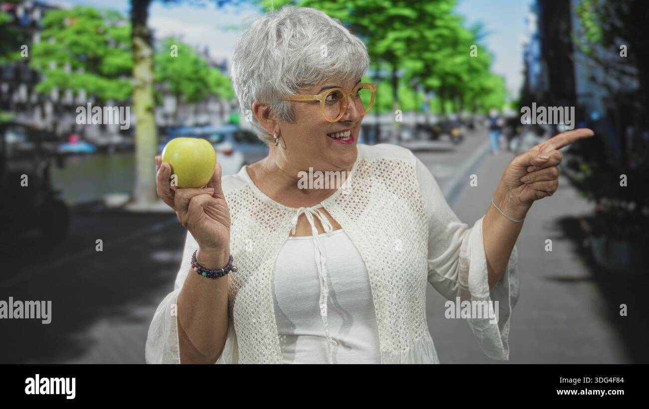 Donna che tiene la mela verde in mano sinistra e punta con la mano destra in strada, sorride con i capelli grigi e indossa gli occhiali; gioiosa curiosità. Foto Stock
