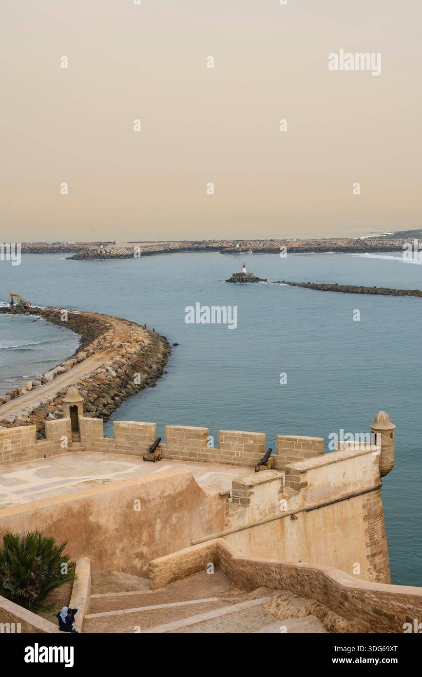 Vista sul forte costiero su una baia tranquilla con un faro distante e muri di pietra. Rabat, Marocco Foto Stock