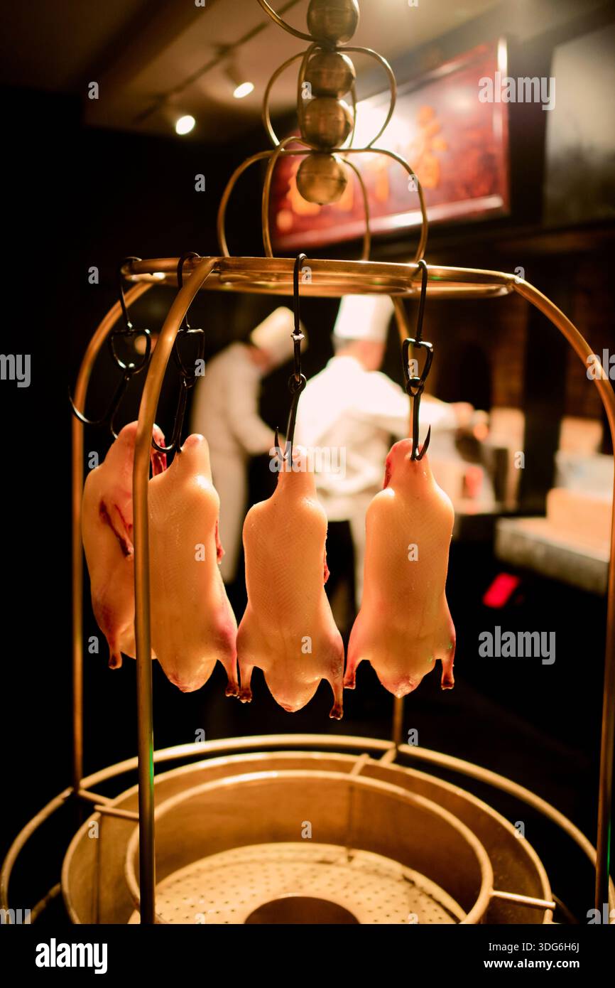 Anatre crude appese ai ganci in una cucina del ristorante con chef che lavorano sullo sfondo. Shanghai, Cina Foto Stock