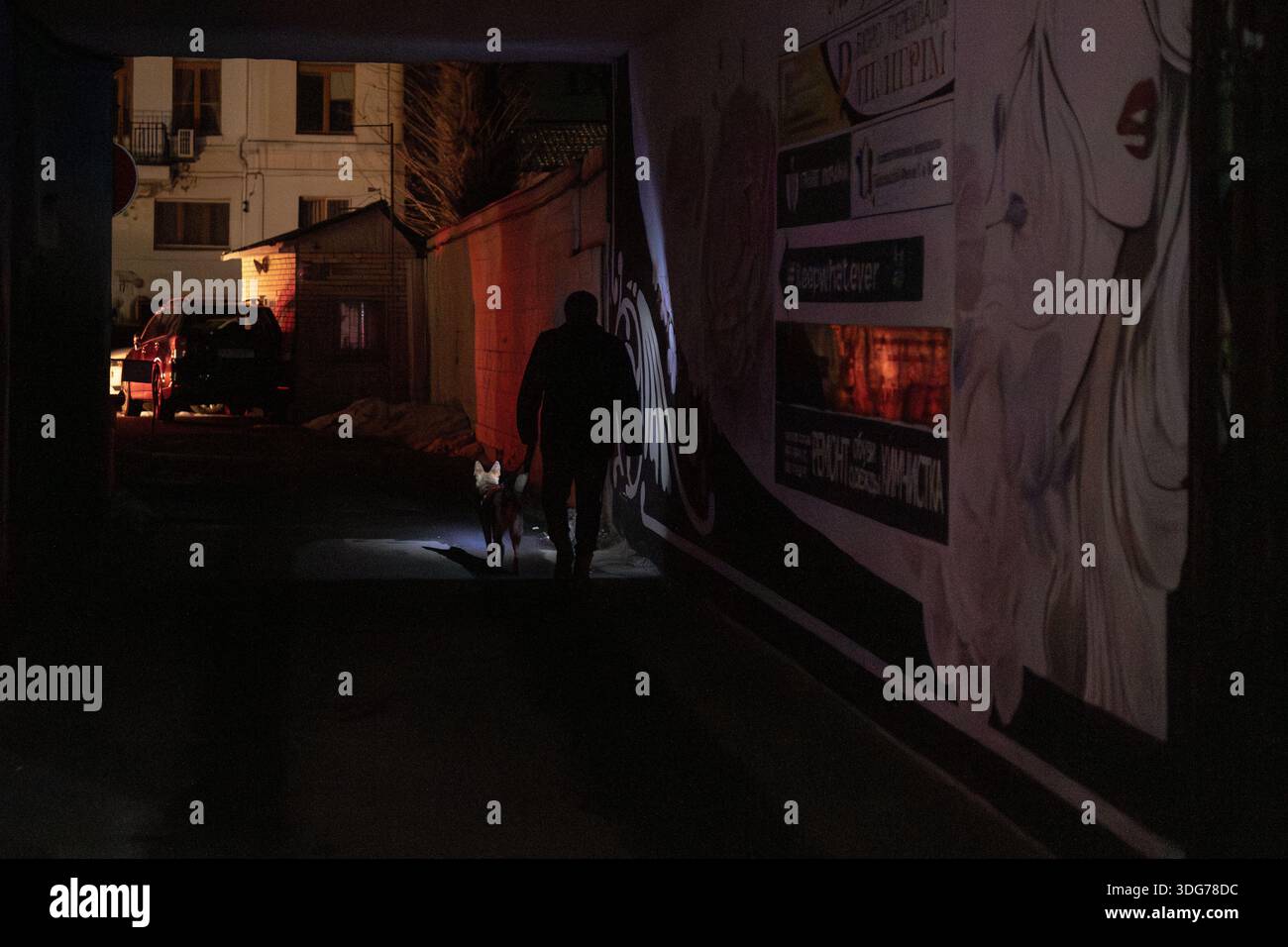 Kiev, Ucraina – 21 dicembre 2022: Una sagoma di un uomo che cammina il suo cane attraverso un arco oscuro su Yaroslaviv Val Street. Solo poche spie illuminano il Foto Stock