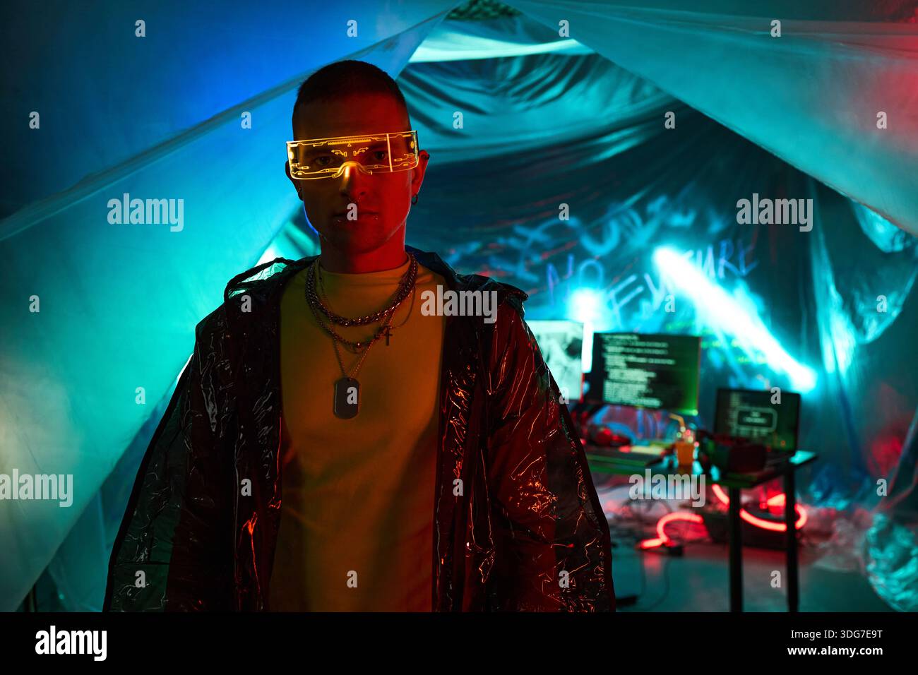 Giovane uomo nero adulto che indossa occhiali futuristici in piedi davanti ai monitor del computer che visualizzano codice in una stanza buia con illuminazione al neon, incarnando il concetto di hacker, non guardando la fotocamera Foto Stock
