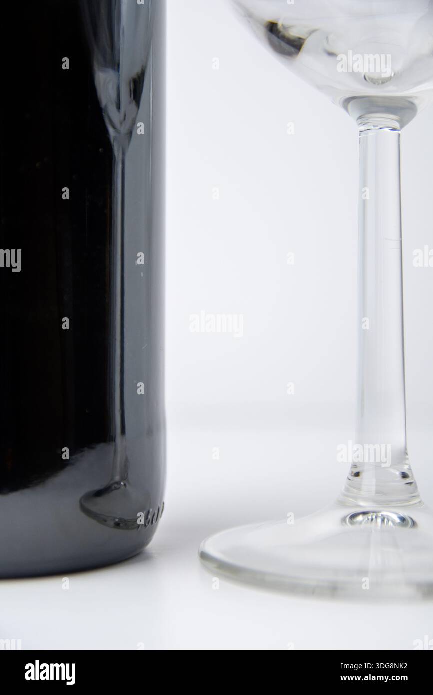 Primo piano minimale di una bottiglia di vino accanto al gambo e alla base di un bicchiere di vino su bianco, che mostra riflessi e uno stile pulito del prodotto. Foto Stock