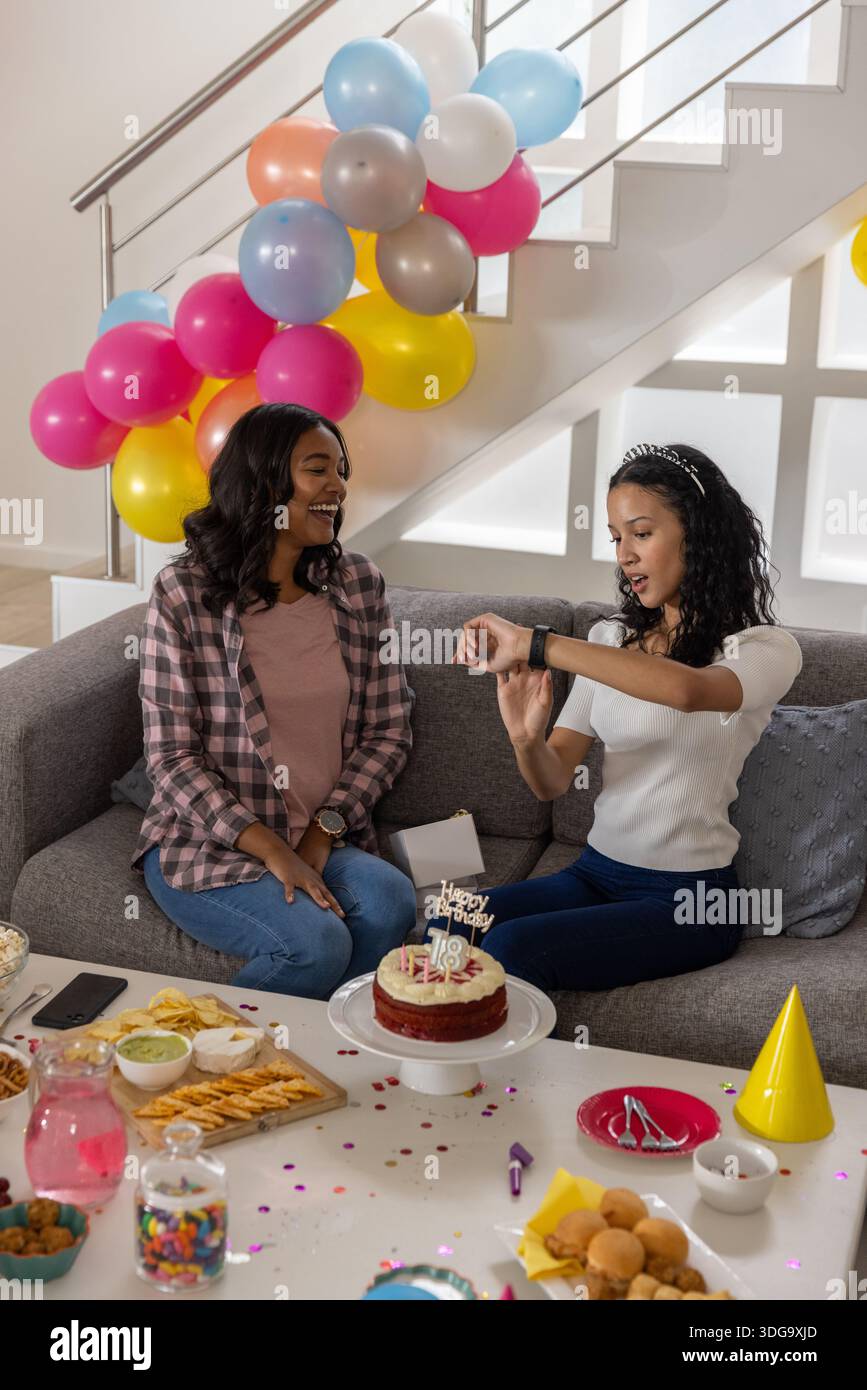 Mamma e figlia adolescente diverse sedute sul divano per festeggiare il diciottesimo compleanno con una torta Foto Stock