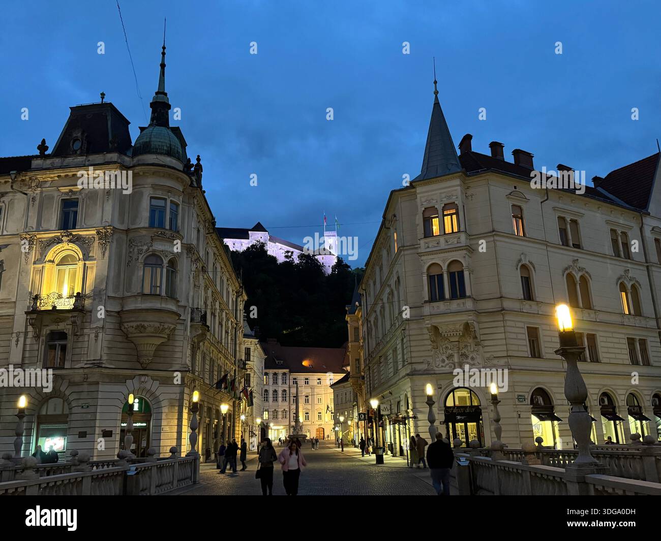 Il castello di Lubiana illuminato al crepuscolo, si affaccia sul vivace lungofiume e sugli edifici storici della capitale slovena - Immagine stock catturata con smartphone