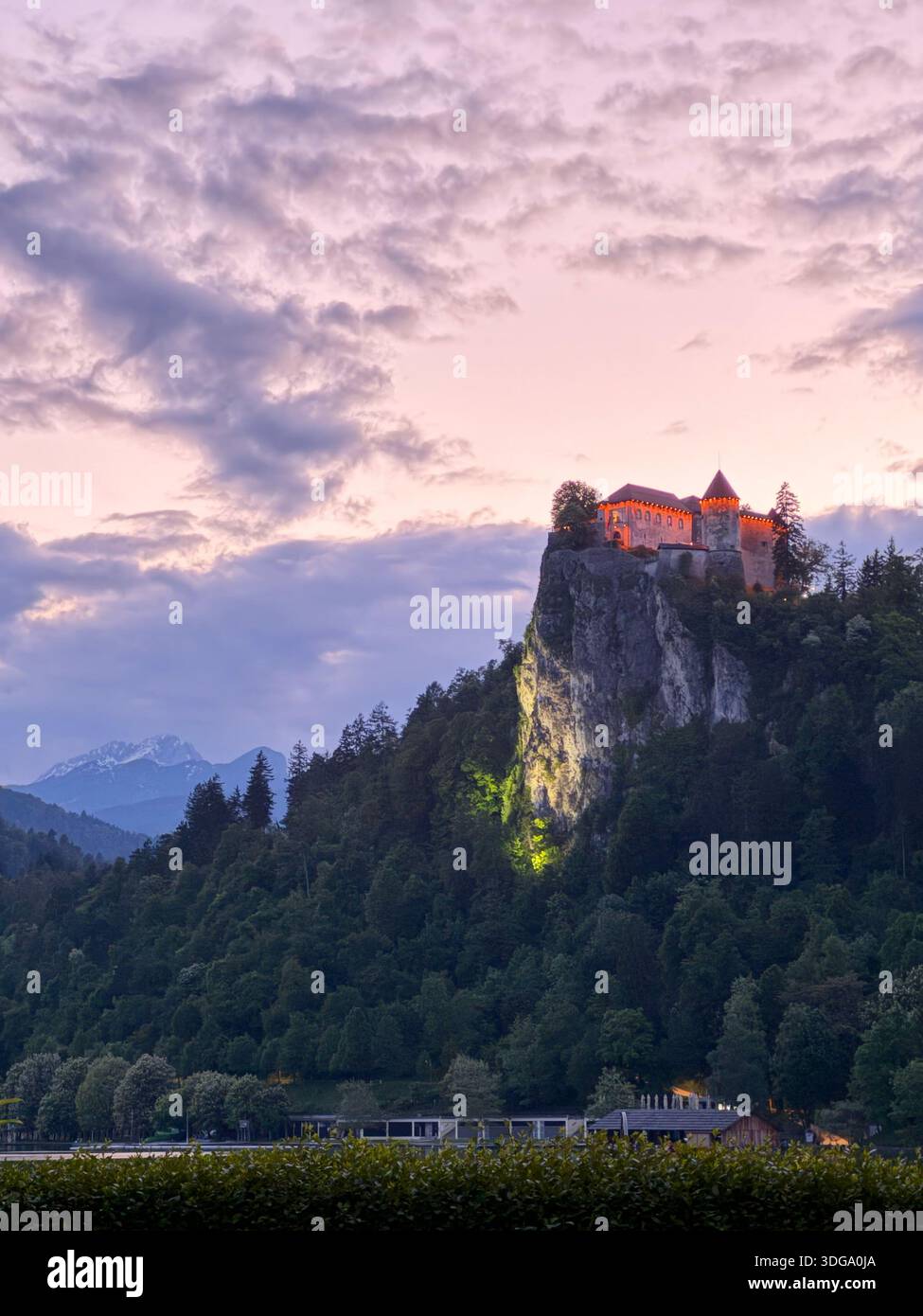 La fortezza illuminata in cima alla collina di Bled si illumina al crepuscolo sopra le scogliere boscose e le vette alpine. Foto Stock