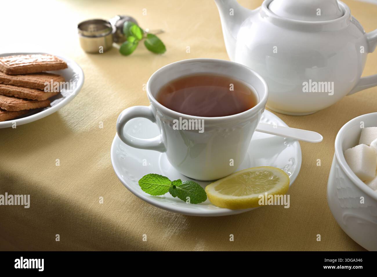 Tè verde in una tazza di ceramica bianca su un piatto con menta e limone su un tavolo con tovaglia marrone con contenitori e biscotti. Vista rialzata. Foto Stock