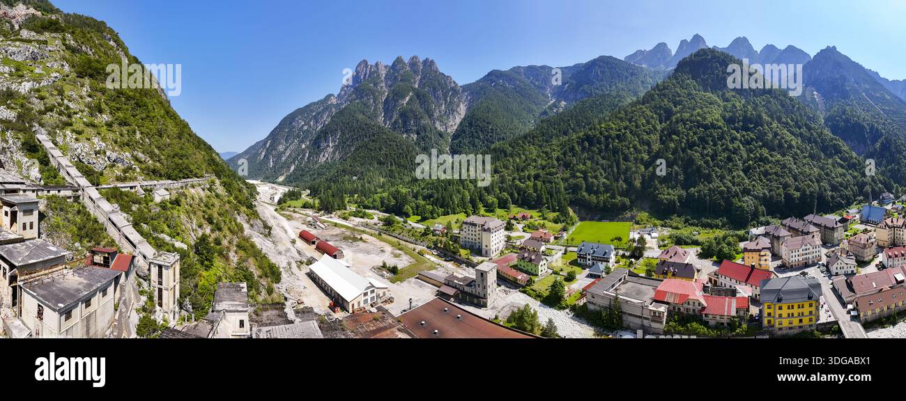 Vista aerea delle aspre montagne incontra una pittoresca cittadina con edifici dal tetto rosso e un antico complesso minerario, Cave del Predil, Friuli-Venezia Giulia, Italia. Foto Stock