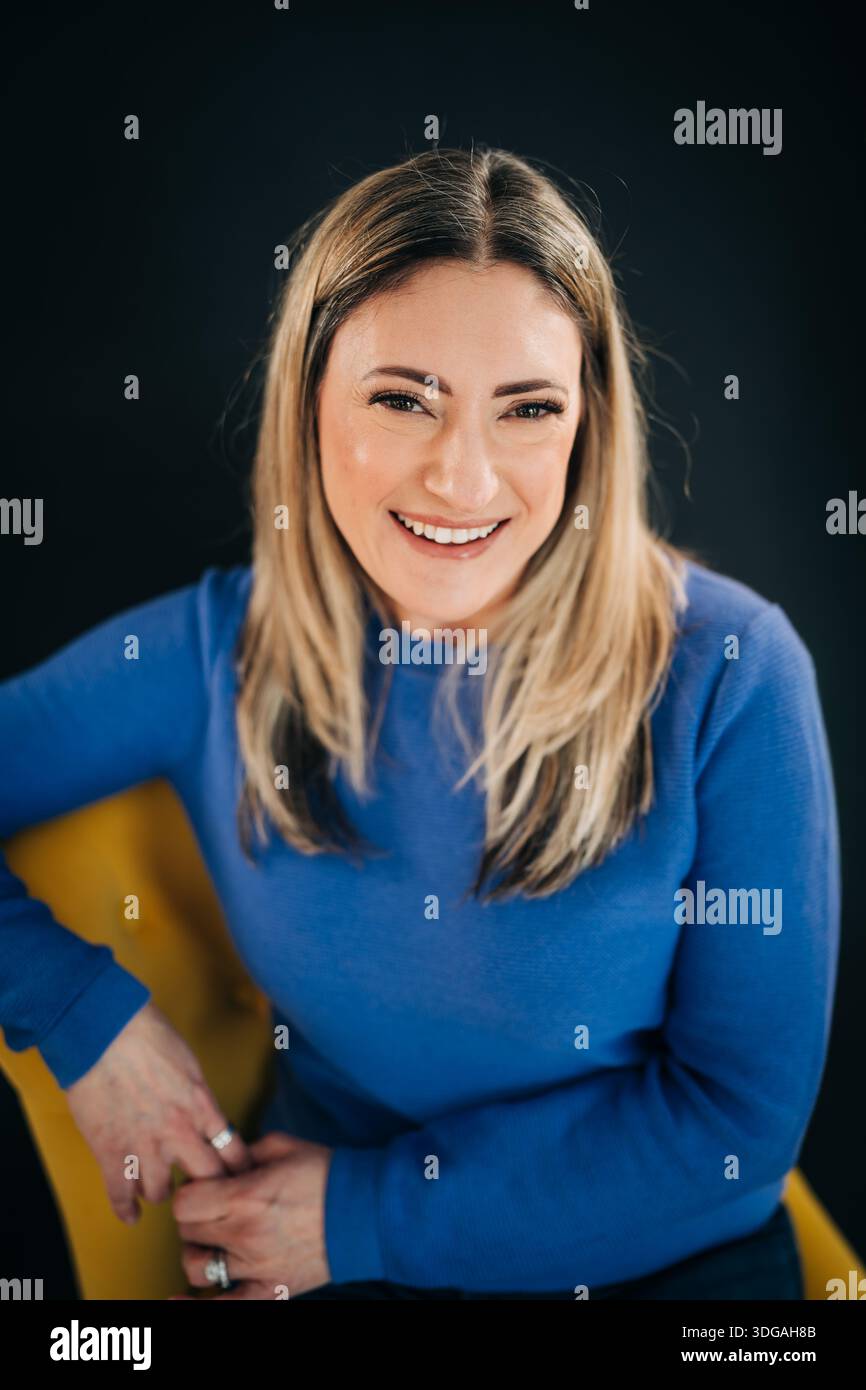 Donna sorridente seduta su una sedia gialla che guarda la macchina fotografica Foto Stock