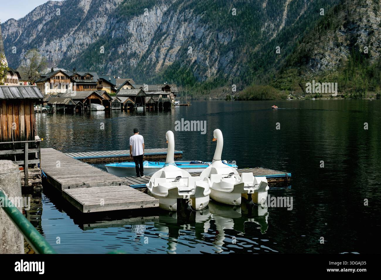 Hallstatt, Austria. Molo in legno con pedalò a cigno ormeggiato presso il lago di montagna. Persona in piedi sul molo del villaggio sul lago con tradizionale boathou di legno Foto Stock