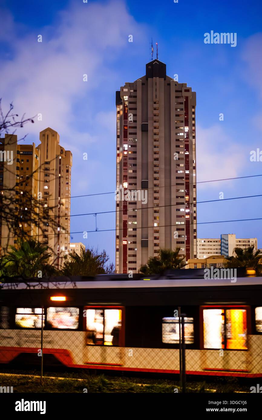 Una vibrante foto notturna di un treno che passa per Benidorm, con il movimento delle auto in contrasto con le torri residenziali statiche e luminose. Foto Stock