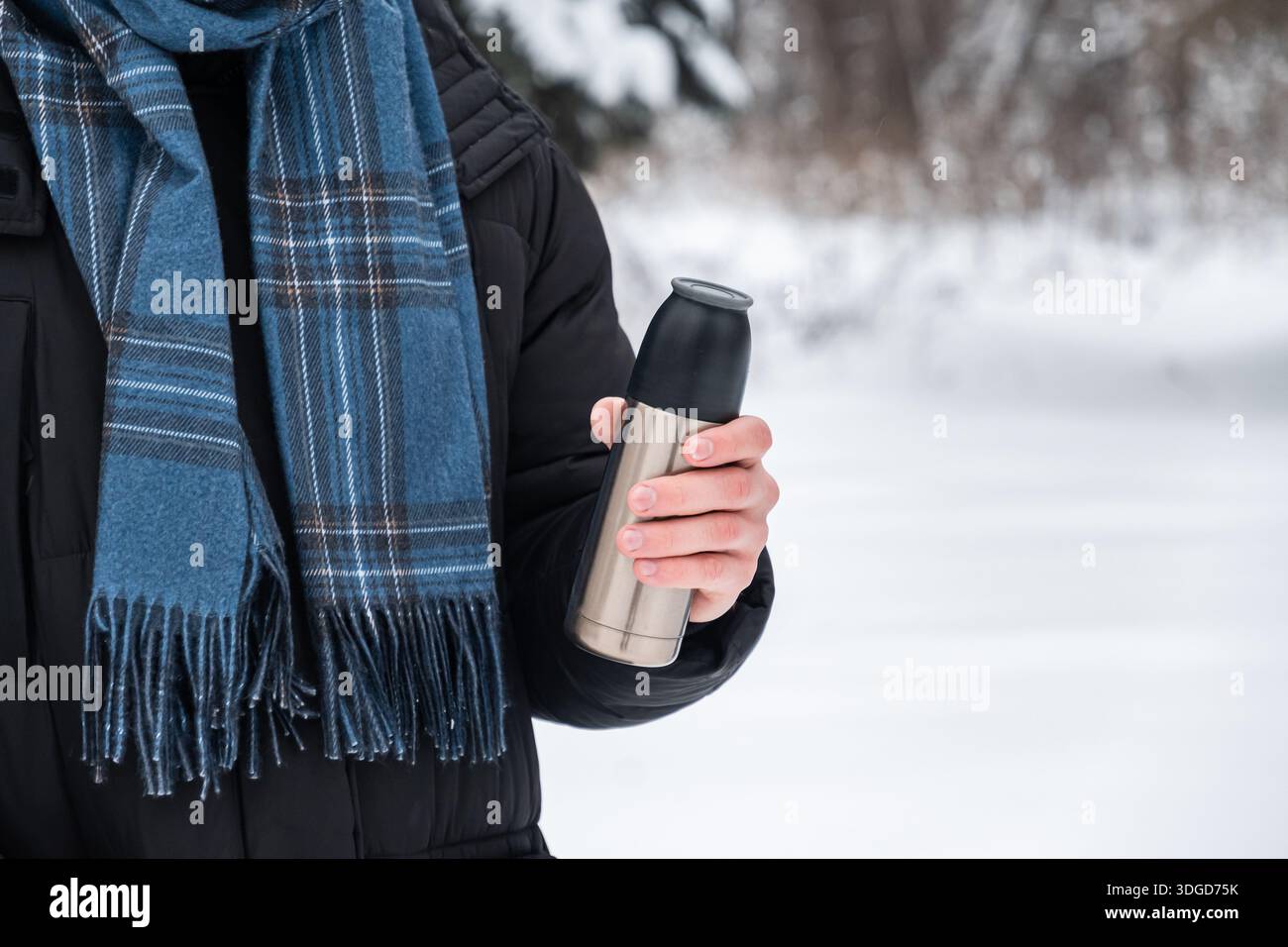 Giovane adulto che tiene in mano termos metallici mentre cammina all'aperto nel parco invernale innevato. Stile di vita in climi freddi, bevande calde e abbigliamento caldo. Foto Stock