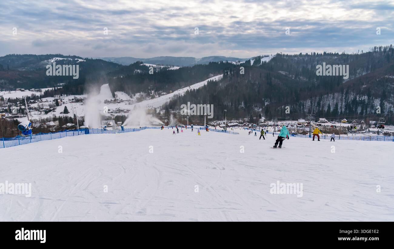 Wisla, Polonia - 6 gennaio 2026: Gli appassionati di sport invernali sciano e snowboard lungo un'ampia pista preparata con cannoni da neve attivi, affacciata su una Foto Stock